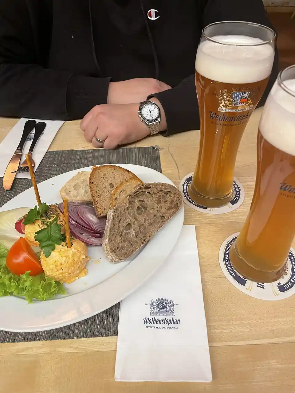 A person wearing a black hoodie with a Champion logo sits at a wooden table with two tall glasses of Weihenstephan beer, a…
