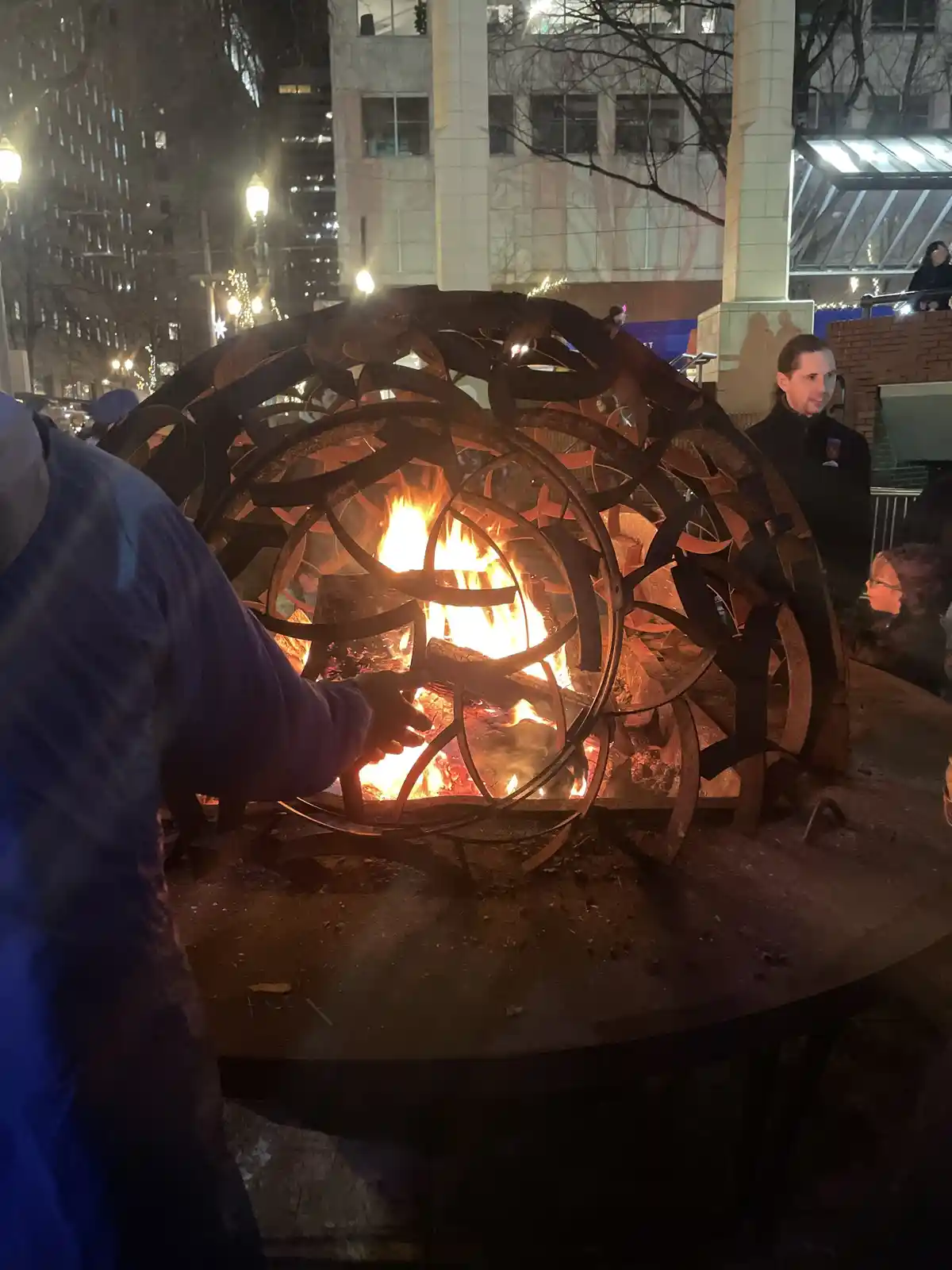 A person in a blue jacket reaches toward a large, ornate metal fire pit with flames inside, while another person stands…