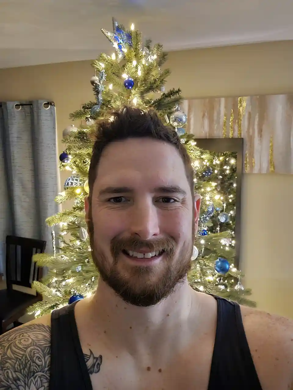 A smiling man with a beard and tattoos wears a black tank top, standing in front of a decorated Christmas tree. The tree…