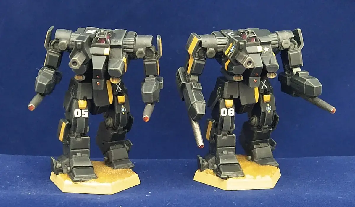 Two identical-looking black and yellow painted model mechs stand side by side on hexagonal bases against a blue background…