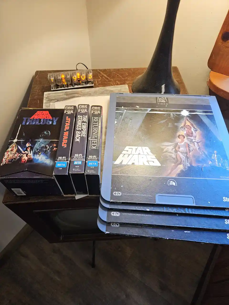 A collection of vintage Star Wars media sits on a wooden table, including a 'Star Wars Trilogy' VHS box set, three…