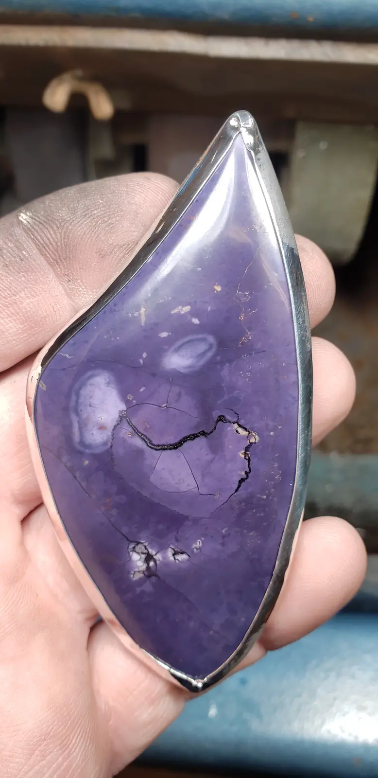 A hand holds a polished, teardrop-shaped purple stone with a prominent dark crack running across its surface, resembling a…