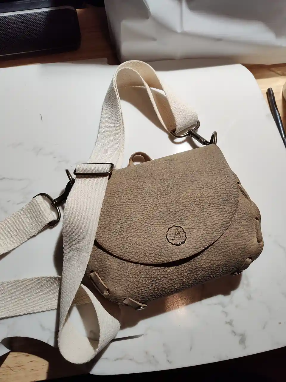 A beige textured leather crossbody bag with a cream canvas strap rests on a white marble surface, featuring a small…