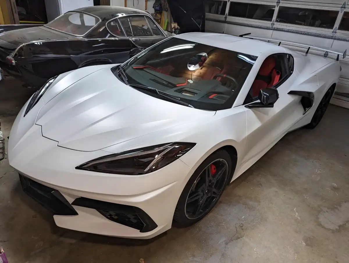 a white matte-finish Chevrolet Corvette C8 sports car with red interior and black wheels is parked in a garage, positioned…