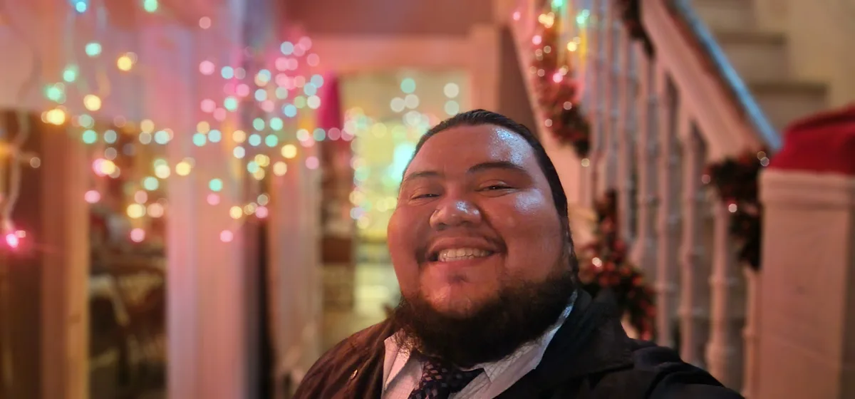 A man with a beard and dark hair, wearing a dark jacket over a collared shirt and tie, smiles broadly with his eyes squinted…