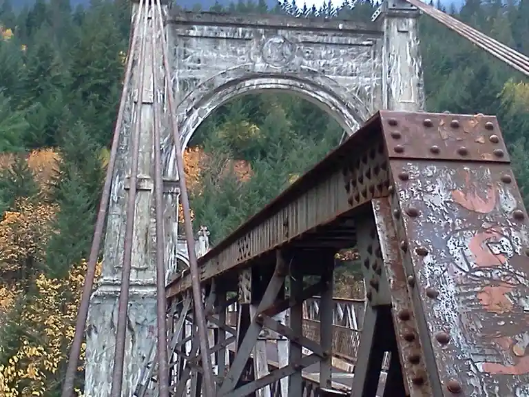 A low-angle shot of a rusty, weathered suspension bridge with ornate stone arches and metal trusses, set against a backdrop…