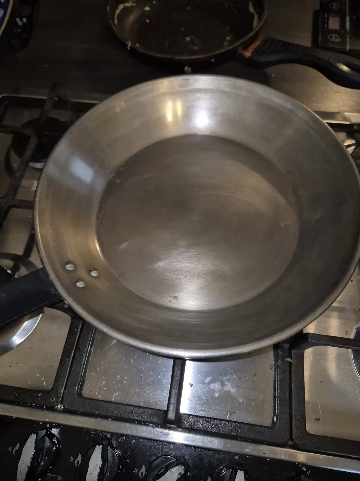 A close-up photo shows a clean, empty stainless steel frying pan sitting on a gas stove burner. The pan has a shiny…