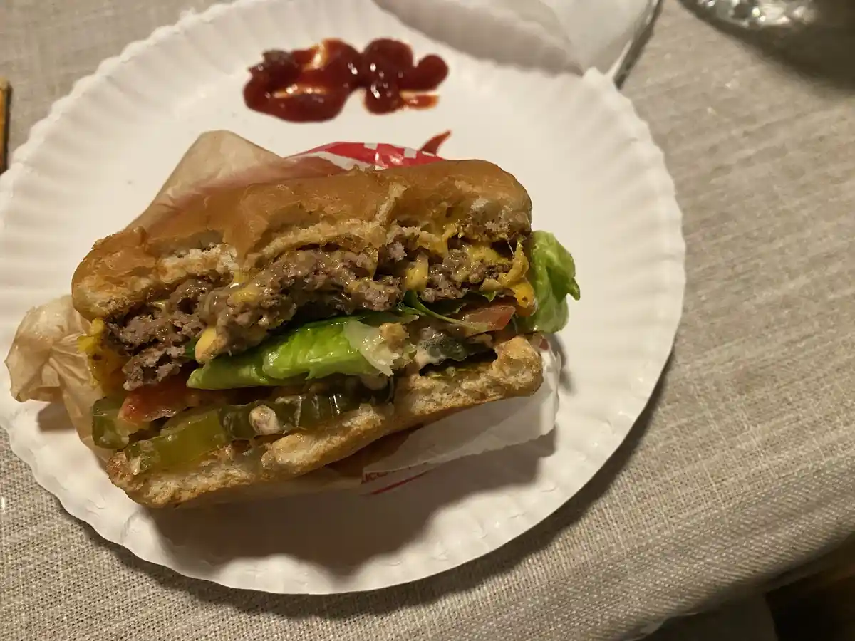 A partially eaten cheeseburger with melted cheese, lettuce, tomato, and pickles sits on a white paper plate with a dollop of…