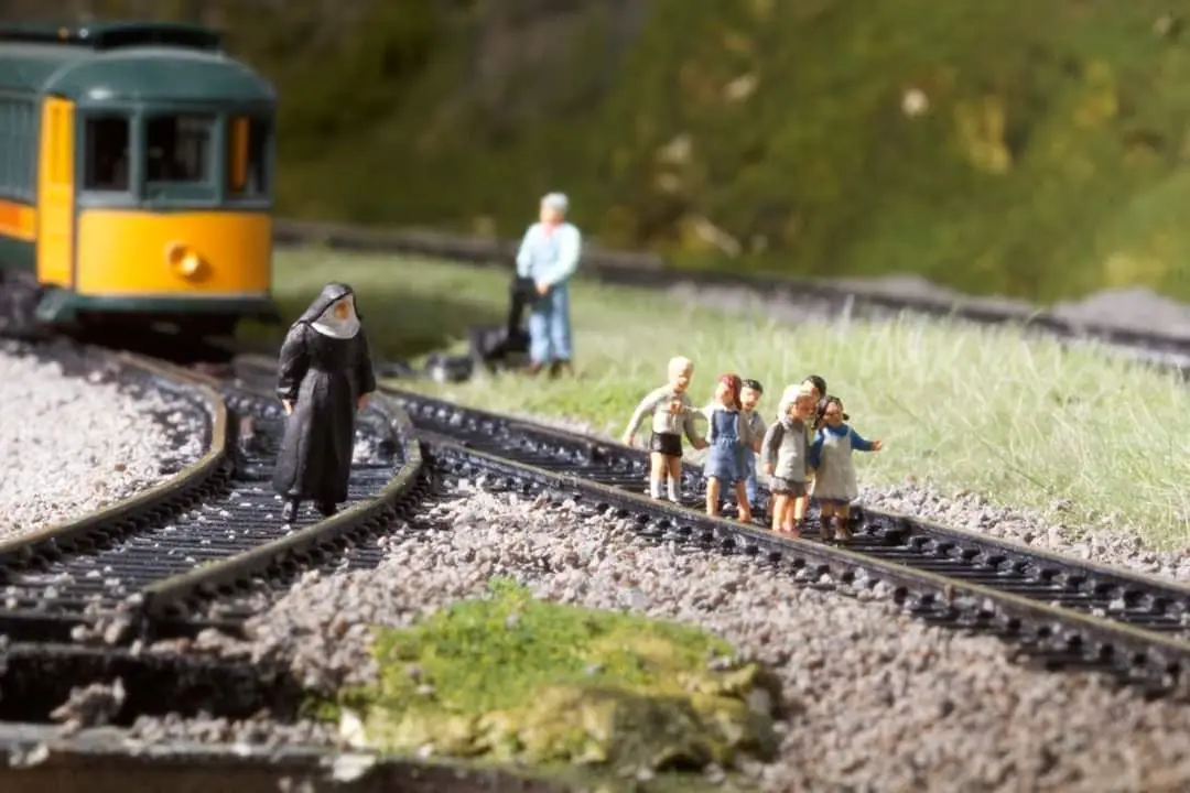 A miniature diorama shows a green and yellow train approaching a set of tracks where a nun stands facing the oncoming train…