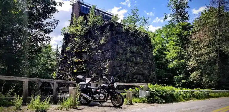A black motorcycle is parked on a roadside next to a weathered stone structure covered in moss and small trees, with a…