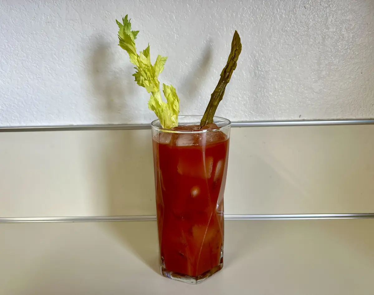 A tall glass filled with a red beverage and ice cubes, garnished with a celery stalk and an asparagus spear, sits on a…