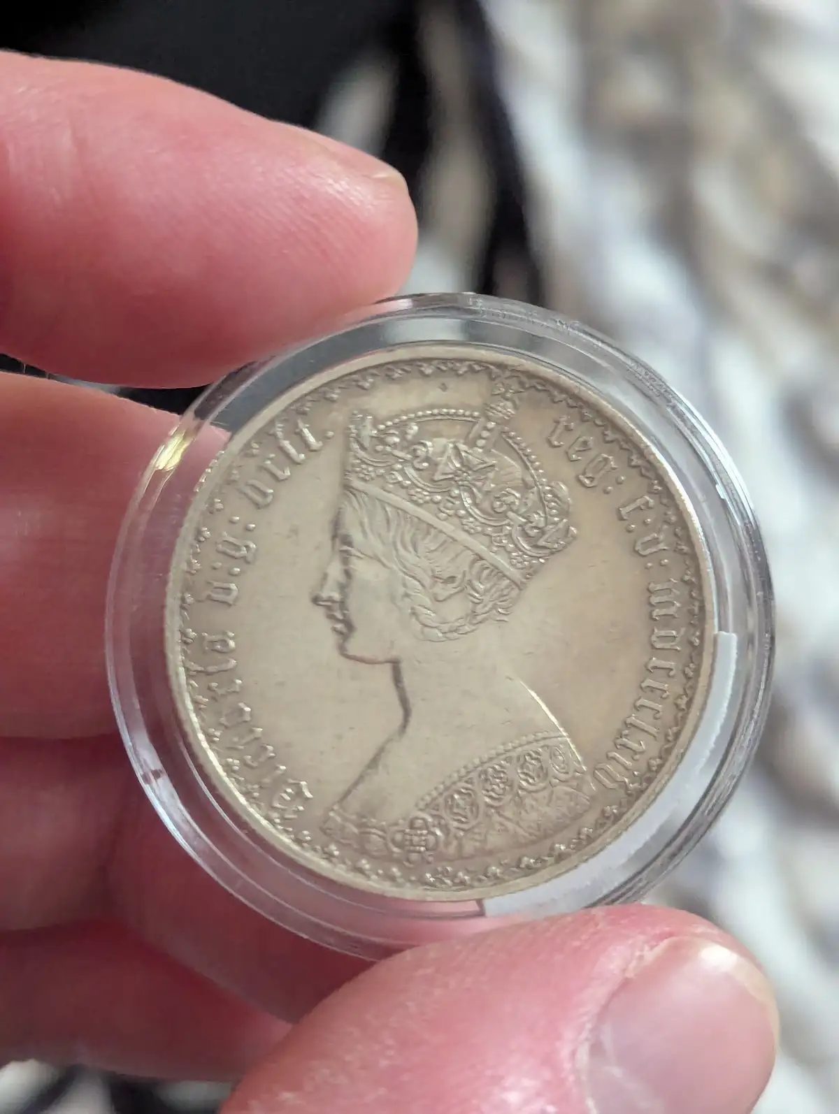 A hand holds a silver coin encased in a clear plastic holder, showing a profile of a crowned woman. The coin's edge has…