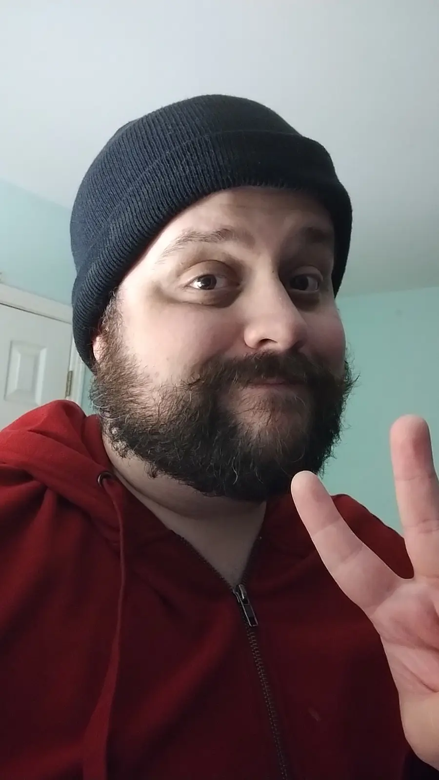 A man with a beard and black beanie wearing a red zip-up hoodie makes a peace sign with his hand while looking at the camera…