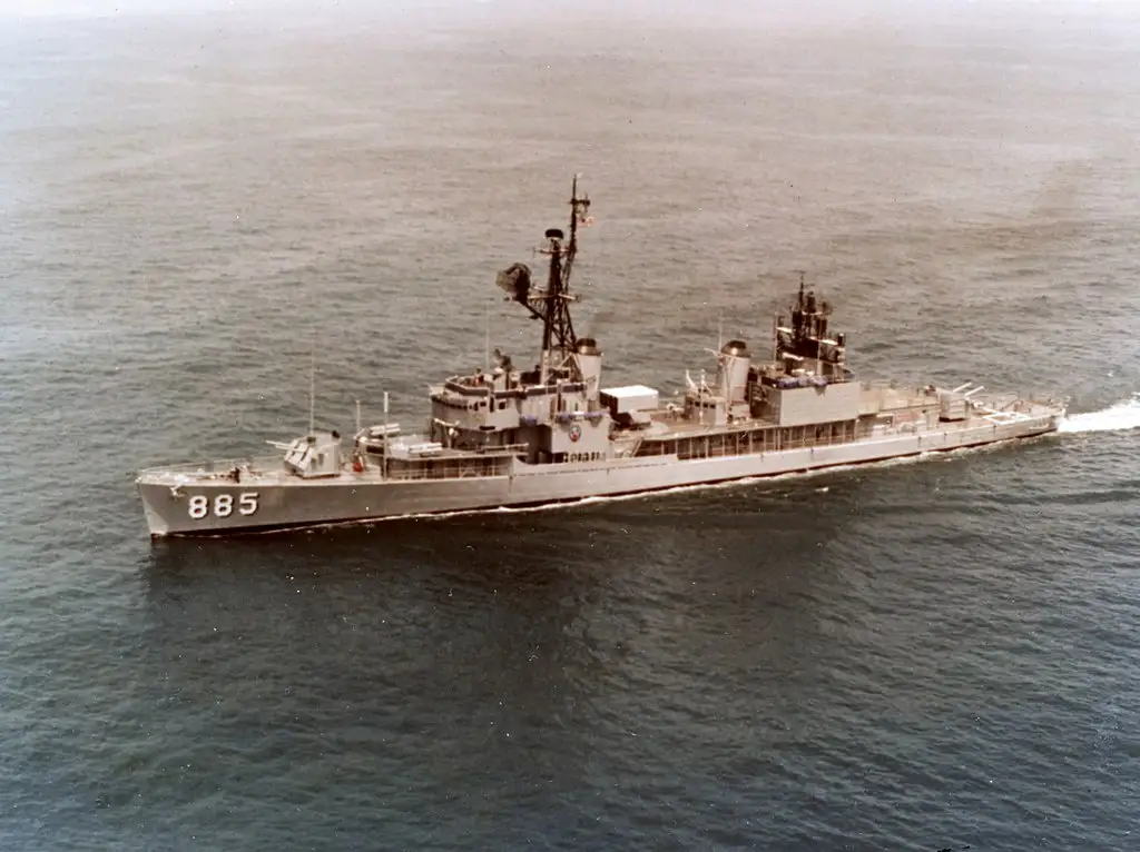 A black and white photograph of a gray naval destroyer sailing on calm ocean waters, its hull number '885' clearly visible…