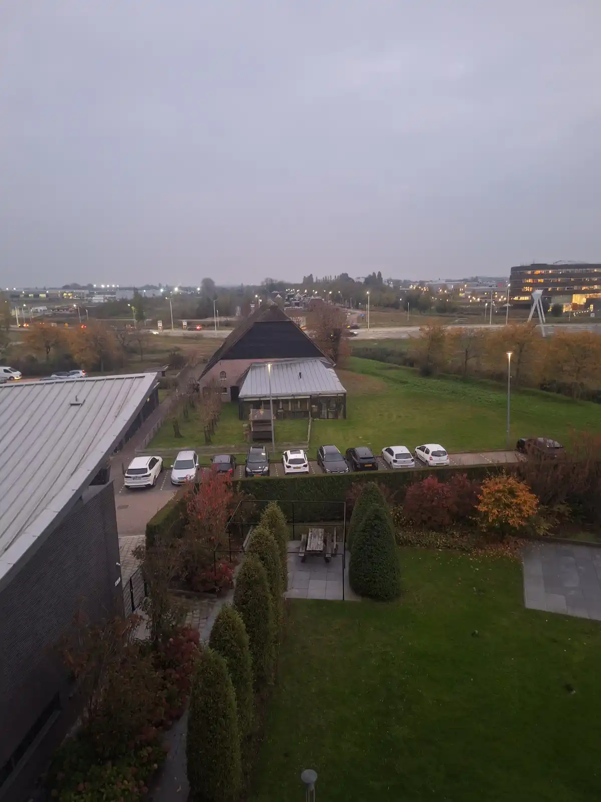 An aerial view of a parking lot with several cars parked in front of a building with a thatched roof and modern extension…