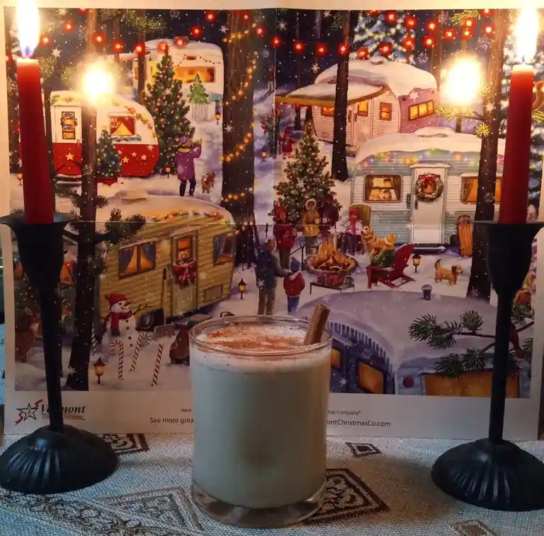 A cozy, festive scene features a glass of eggnog with a cinnamon stick and nutmeg garnish in the foreground, flanked by two…