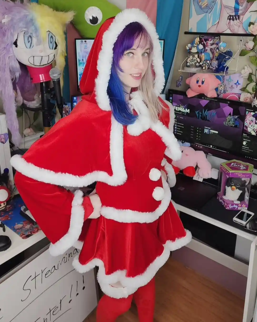 A person with purple and blue hair wears a red, white-furred Santa-style hooded outfit while posing in a room filled with…