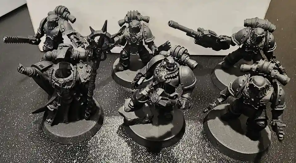 A black-and-white photo shows six heavily armored, grim-faced miniature warriors in battle poses, each on a circular base…