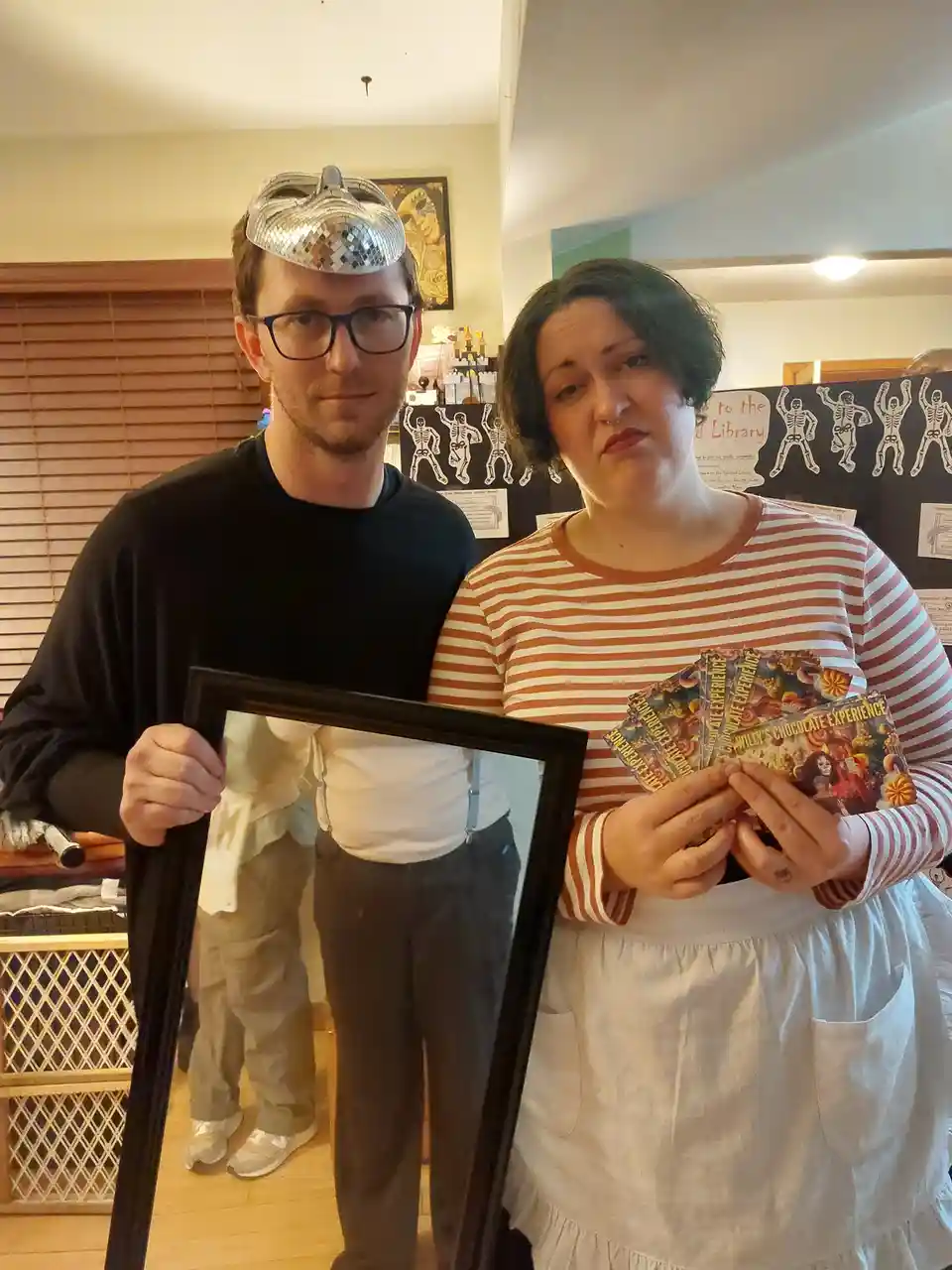 A man wearing a disco ball mask and glasses holds a mirror, while a woman in a striped shirt and apron holds tickets to…