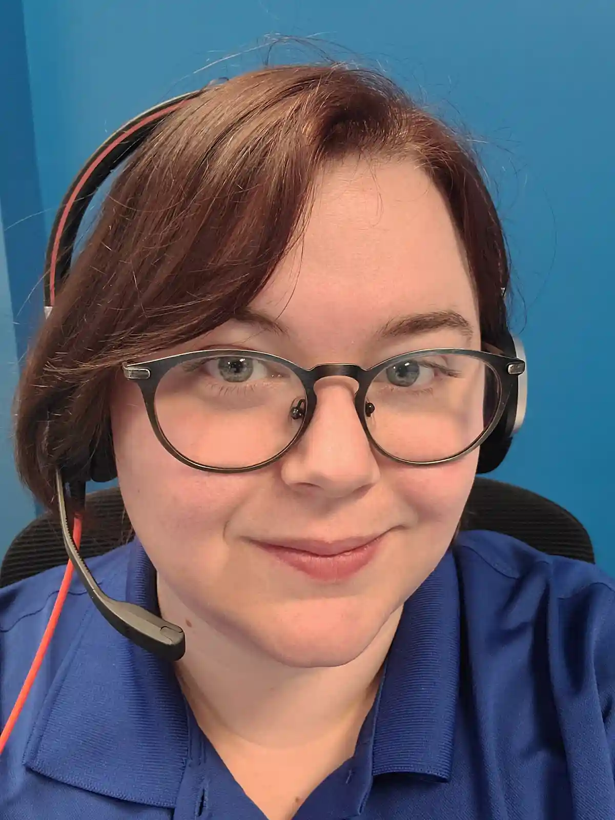 A person with brown hair, wearing glasses and a headset with a microphone, smiles slightly while looking at the camera. They…