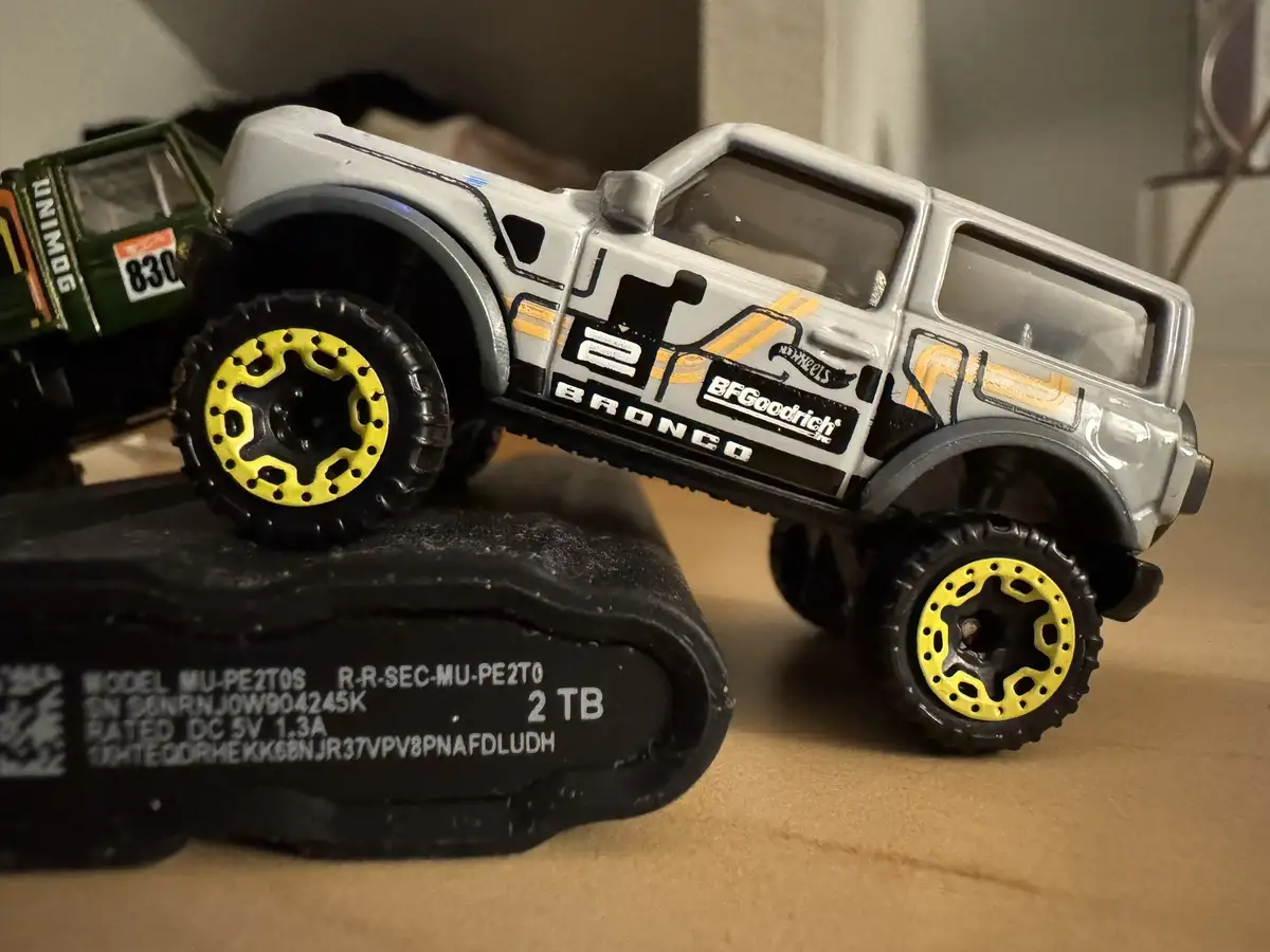 A silver Hot Wheels Broncos toy truck with yellow wheels is posed on top of a black USB flash drive labeled '2 TB'. The…