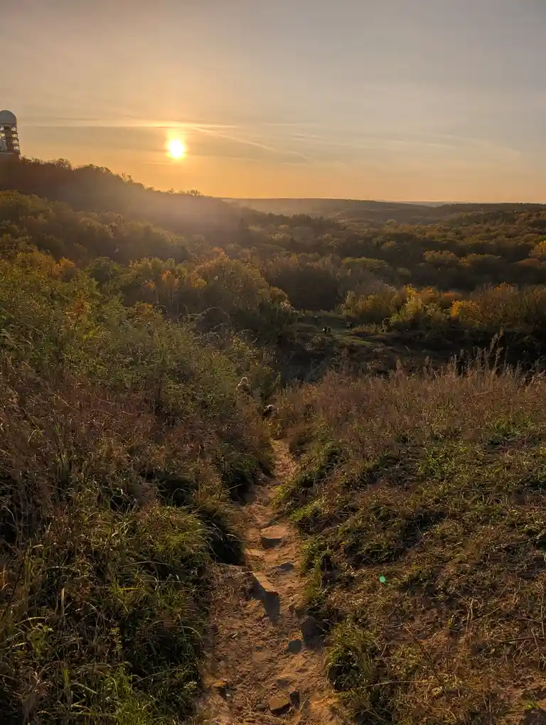 A scenic photo of a dirt hiking trail winding through a forest at sunset, with the sun low in the sky casting warm light…