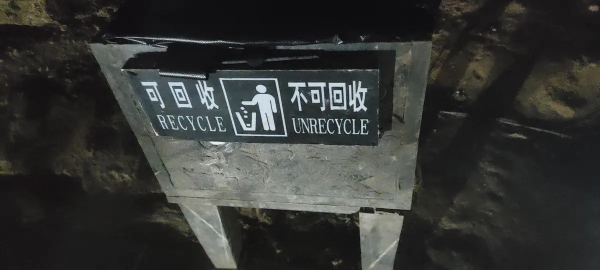 A dark, weathered recycling bin sign with Chinese characters and English text. The left side reads '可回收' (recyclable) and…