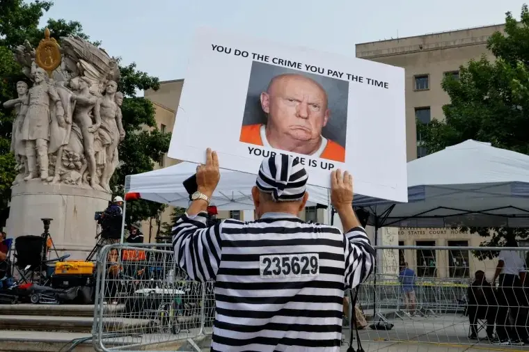 A person in a black-and-white striped prisoner uniform with the number 235629 on their back holds up a large white sign. The…