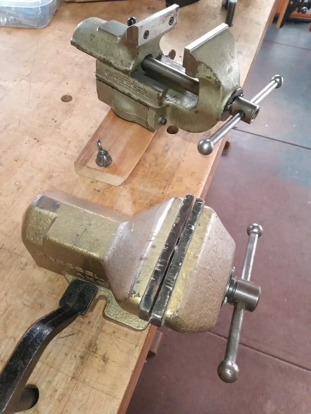 Two worn, brass-colored Gressel brand bench vices sit on a wooden workbench, one with its jaws open and the other closed…