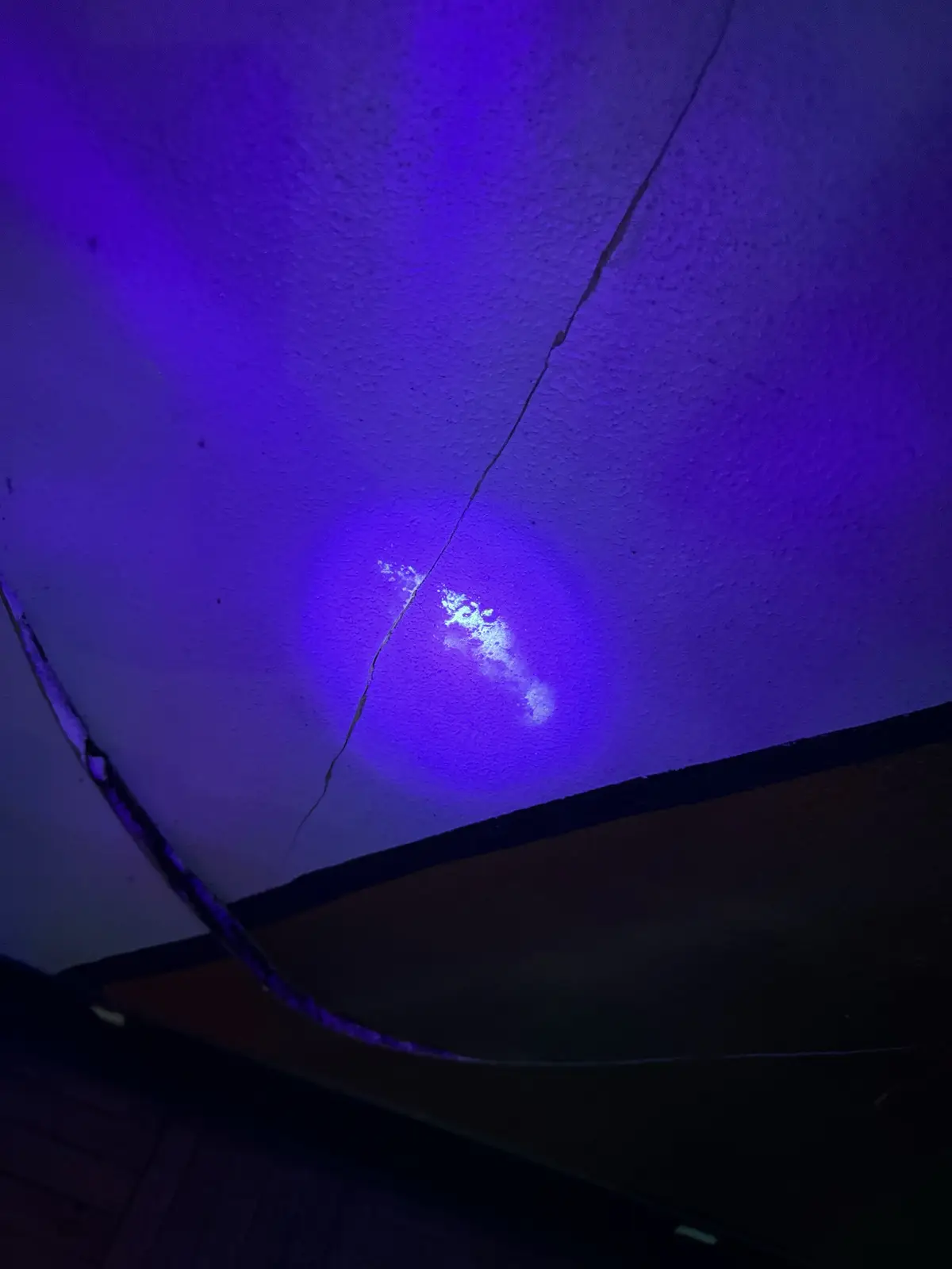 A close-up photo shows a textured ceiling bathed in purple light, with a prominent crack running diagonally. A bright…