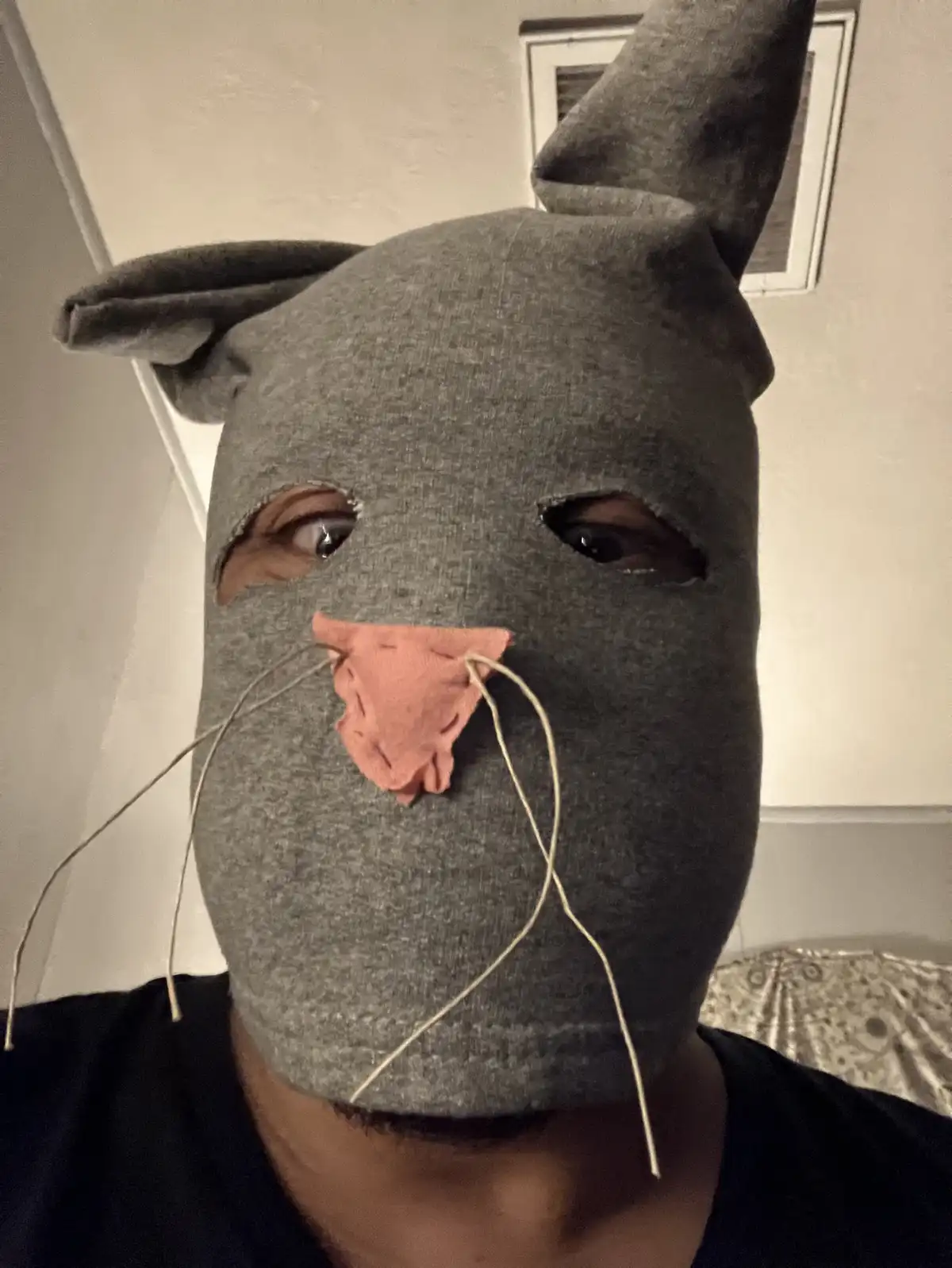 A person wears a homemade grey fabric mask resembling a rat, complete with ears, a pink stitched nose, and string whiskers…