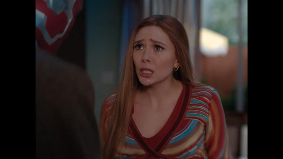 A woman with long brown hair and a striped top looks horrified and confused while facing a partially visible person in a red…