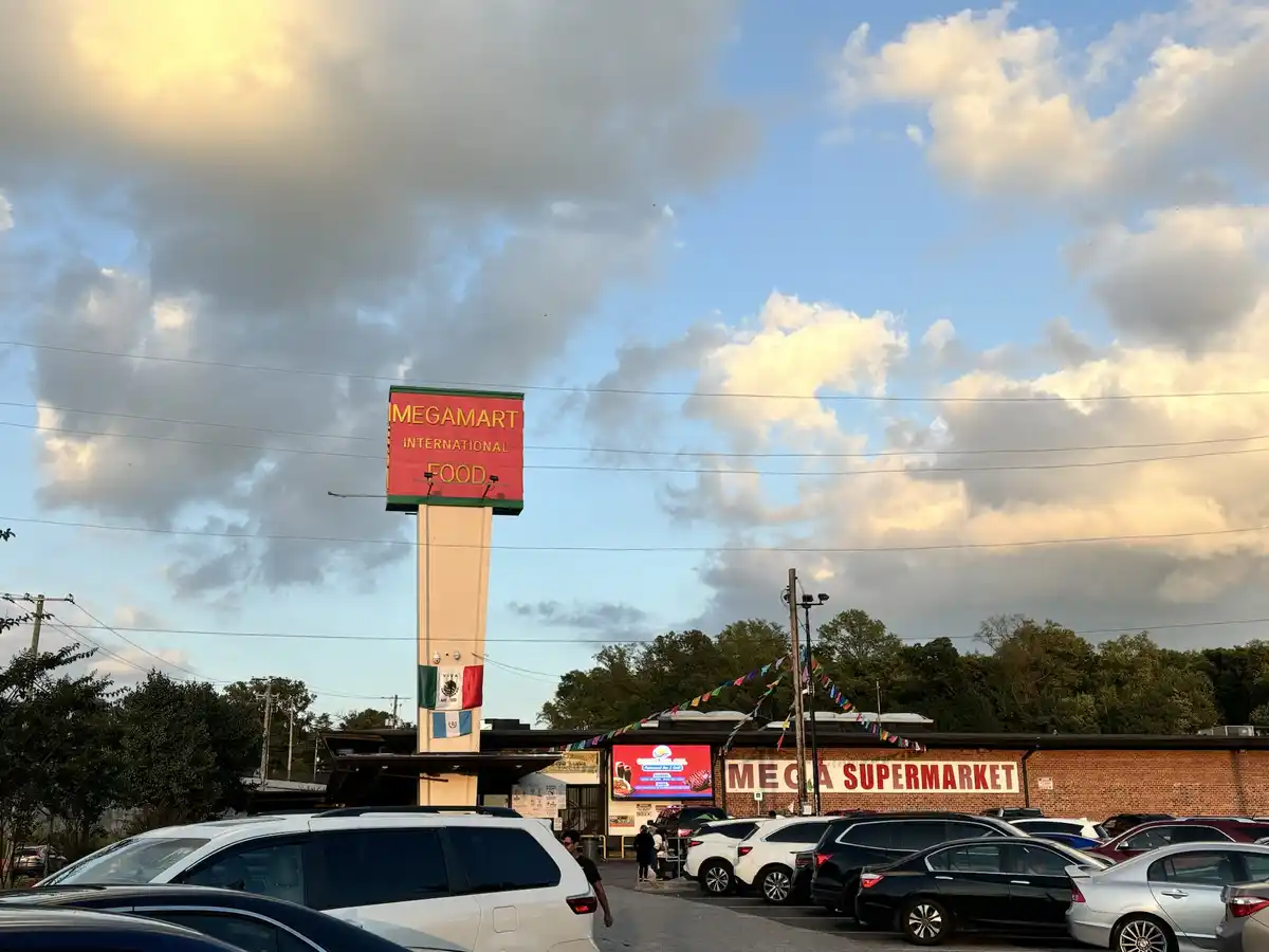 An exterior photo of a supermarket named 'Megamart International Food' with a tall red sign, parked cars, and a partly…