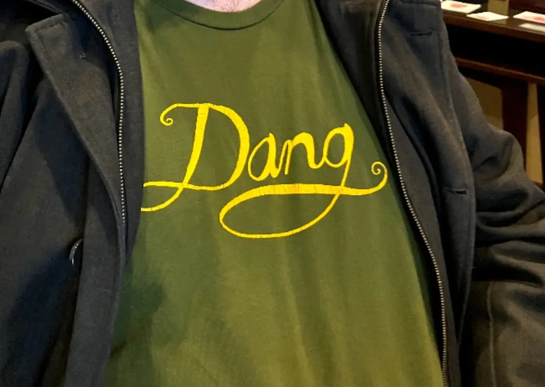 A close-up photo shows a person wearing a green t-shirt with the word 'Dang' printed in large, yellow, cursive script across…