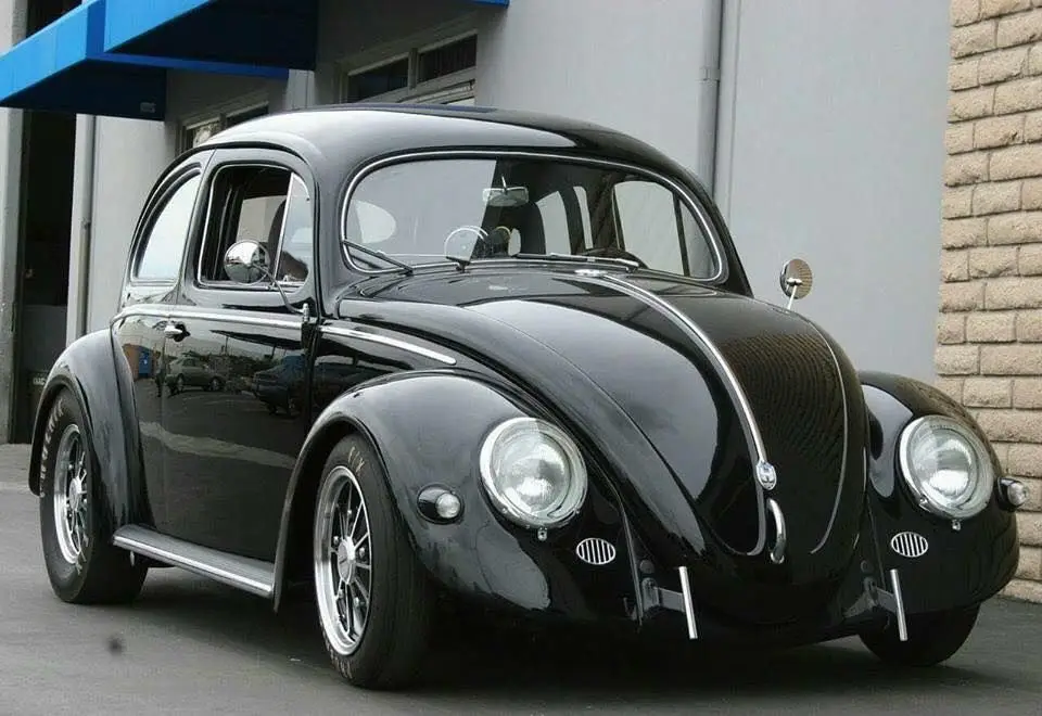 A glossy black vintage Volkswagen Beetle is parked outside a building with a blue awning and brick wall. The car has chrome…