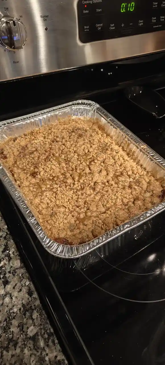 A rectangular aluminum baking pan filled with a golden-brown crumble dessert sits on a black electric stovetop. Above it…