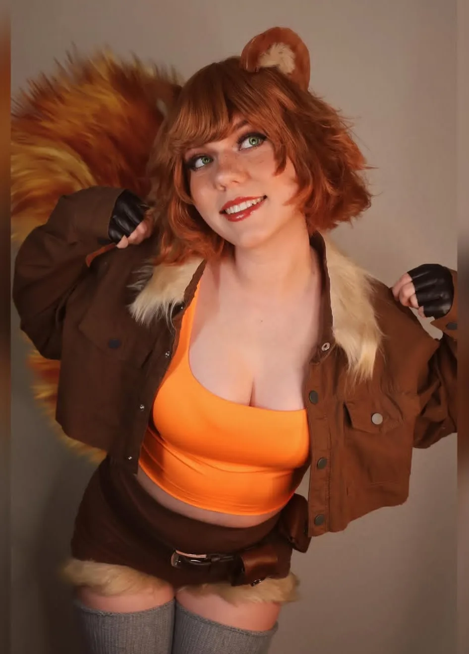 A person in a detailed squirrel cosplay, featuring orange-brown fur, a large bushy tail, and ears, poses confidently in a…