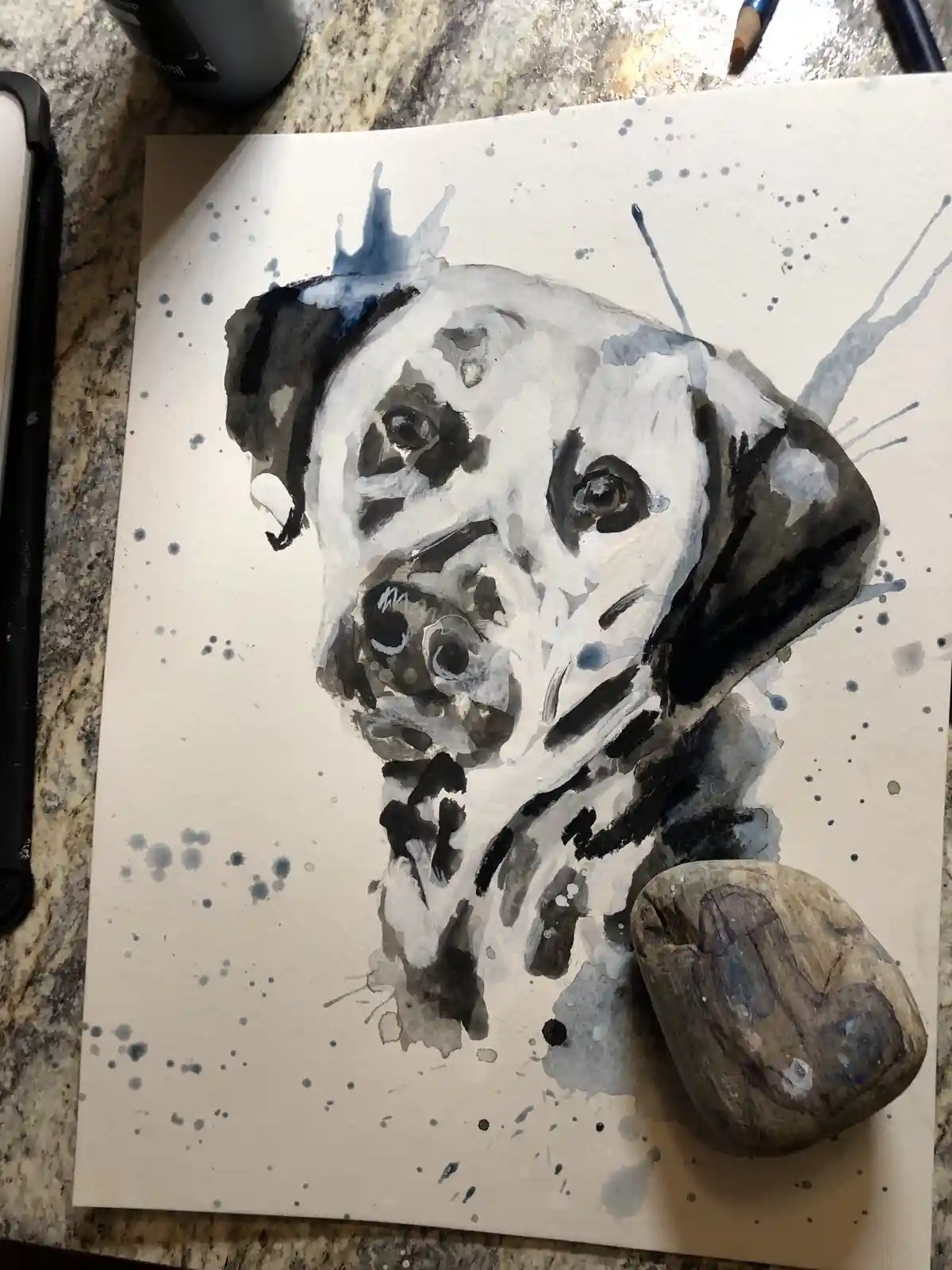 A watercolor painting of a dog's head on paper, surrounded by blue paint splatters, with a small, painted rock placed beside…