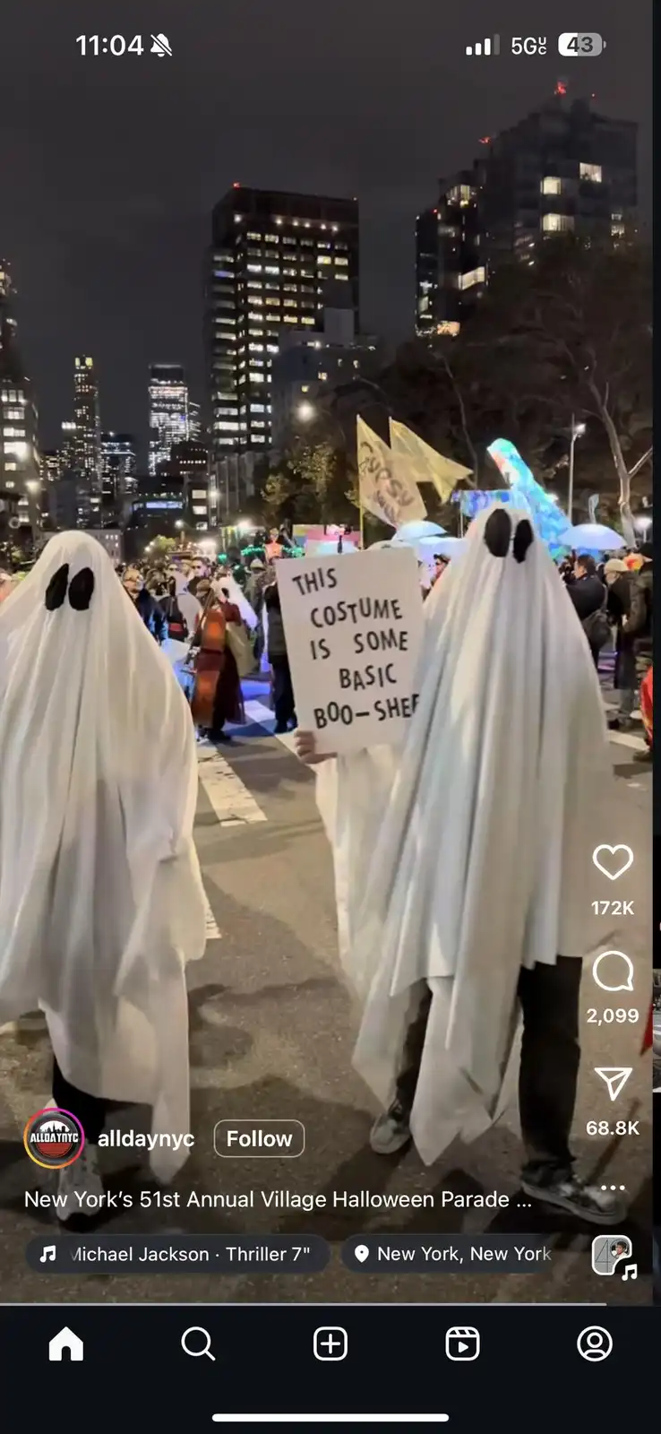 Two people dressed as ghosts with simple black dot eyes walk in a nighttime Halloween parade, holding a sign that reads…
