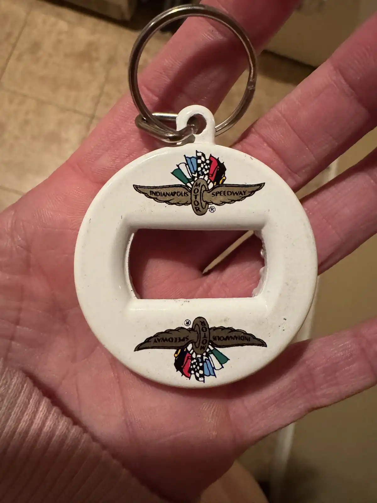 A hand holds a white circular bottle opener keychain with the Indianapolis Motor Speedway logo printed on both top and…