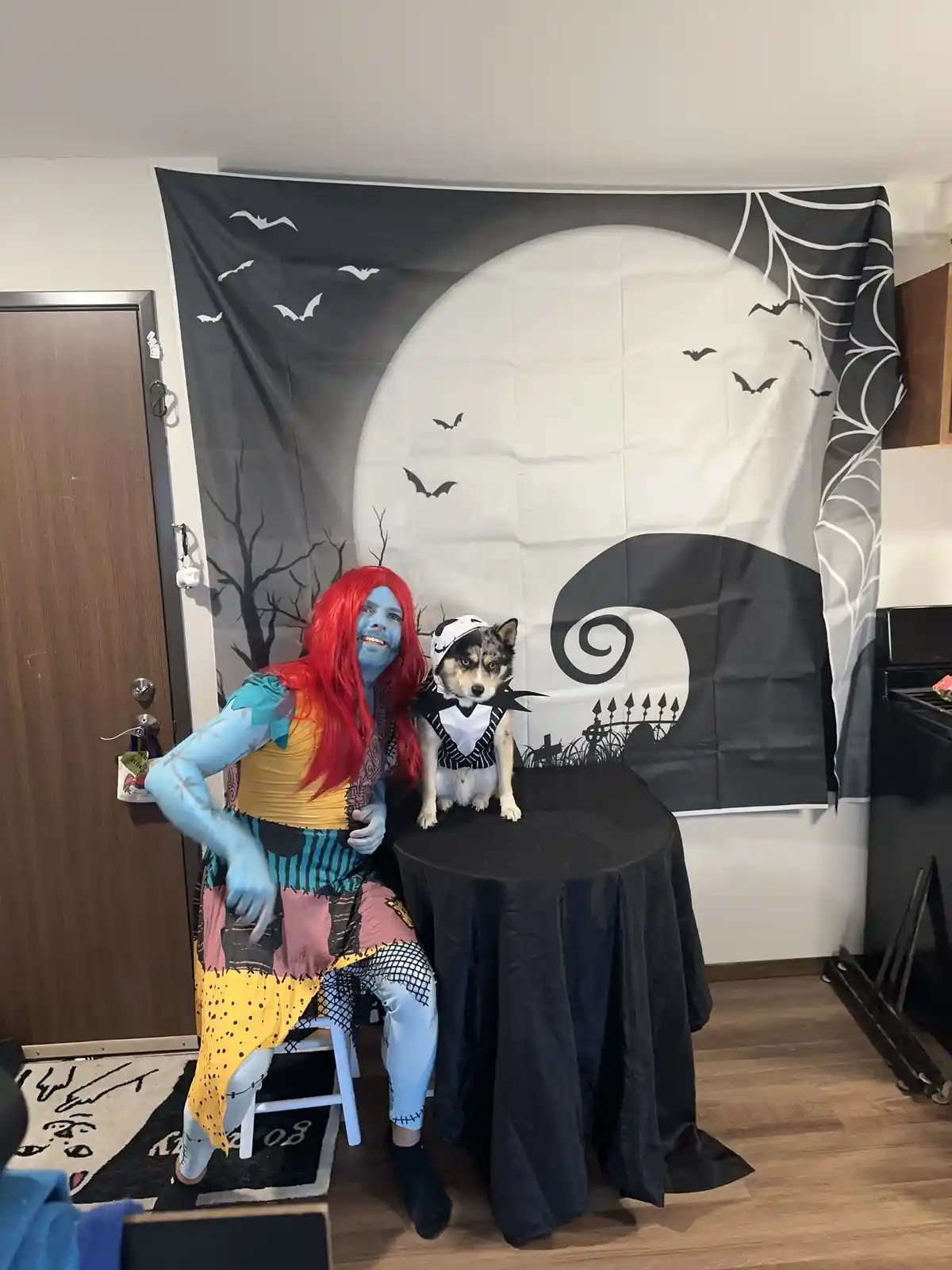 A person with blue face paint, red wig, and a patchwork costume poses beside a dog dressed as Jack Skellington, both in…
