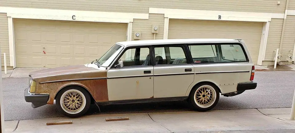 A vintage white station wagon with significant rust on its front half is parked in front of beige garage doors labeled 'C'…