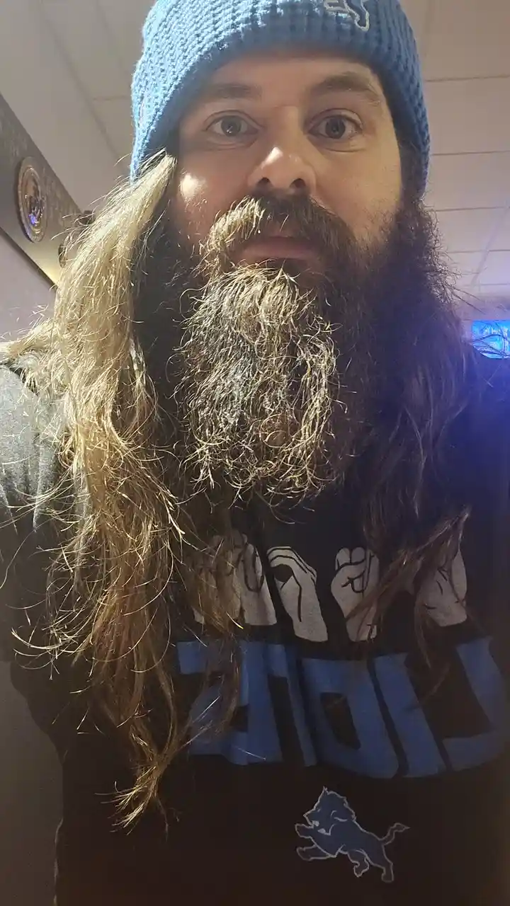 A man with a long, thick beard and long hair wears a blue Detroit Lions beanie and a matching Lions t-shirt. He stares…