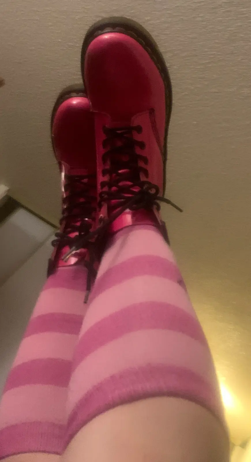 A close-up, low-angle shot shows a person's legs wearing bright pink, shiny combat boots with black laces and…