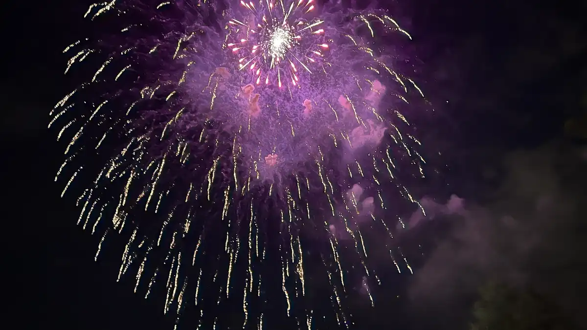 A vibrant purple and gold fireworks explosion fills the night sky, with smoke trailing downward. The overlaid text reads…