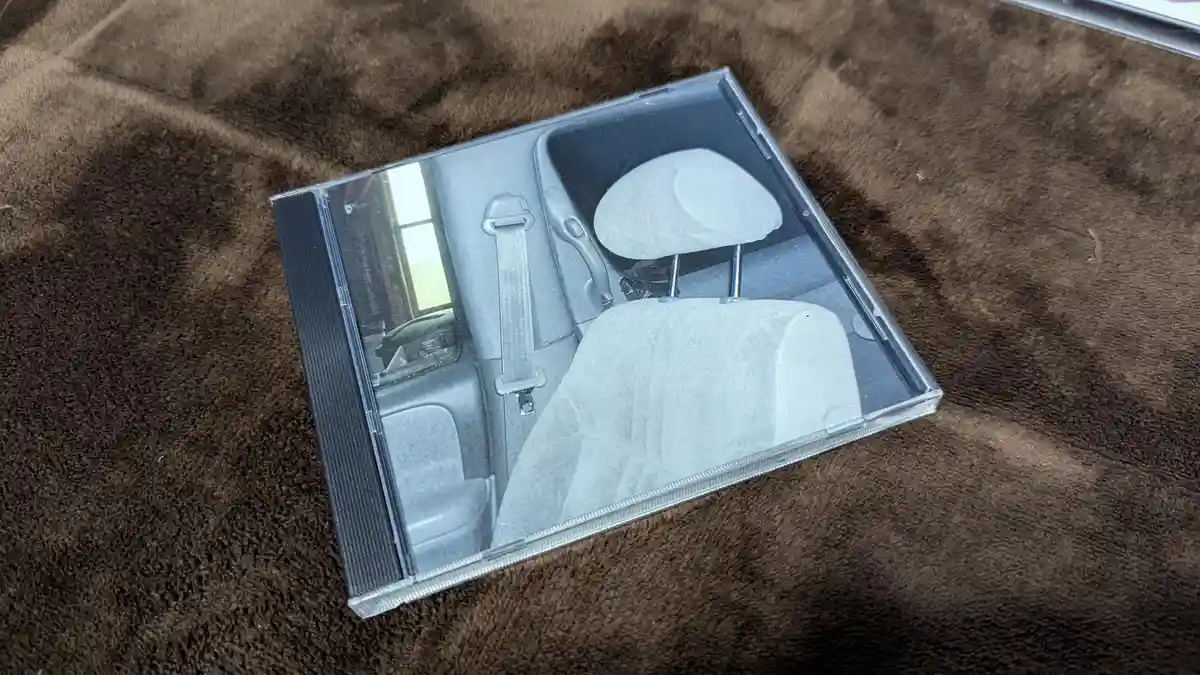 A clear plastic CD case rests on a brown textured blanket, its cover displaying a black-and-white photo of a car's interior…