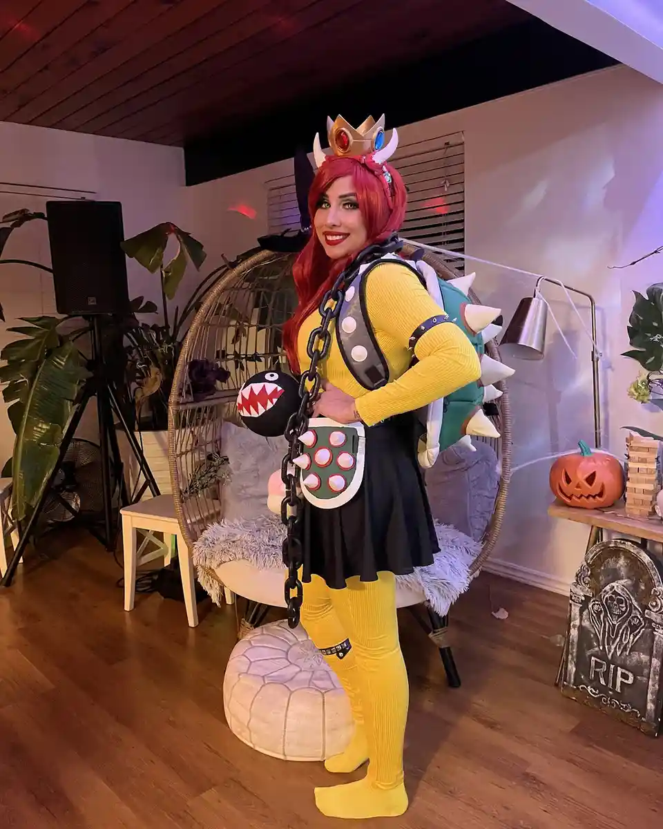 A person in a detailed Bowser Jr. costume from Super Mario, complete with crown, spiked shell, and chain, poses indoors with…
