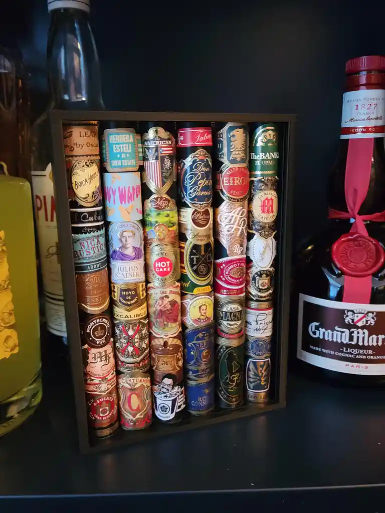 A dark wooden display case holds numerous cigars standing upright, their colorful, ornate bands resembling book spines…