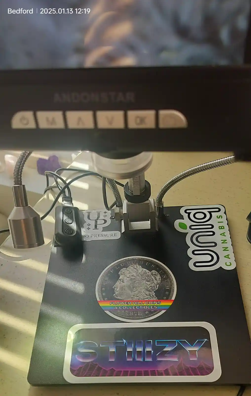 A close-up photo shows a coin with a rainbow sticker reading 'FLOWER HILL COINS & COLLECTIBLES' placed on a black device…