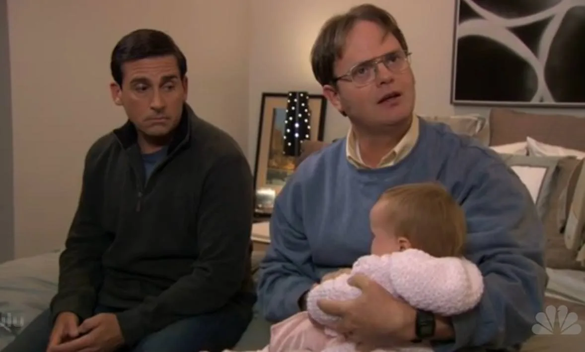 Two men sit on a bed; the man on the right holds a baby wrapped in pink and looks upward with a surprised expression, while…